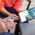 Man using an Alanté finger glucose tester with a women holding a tablet to show Alanté website